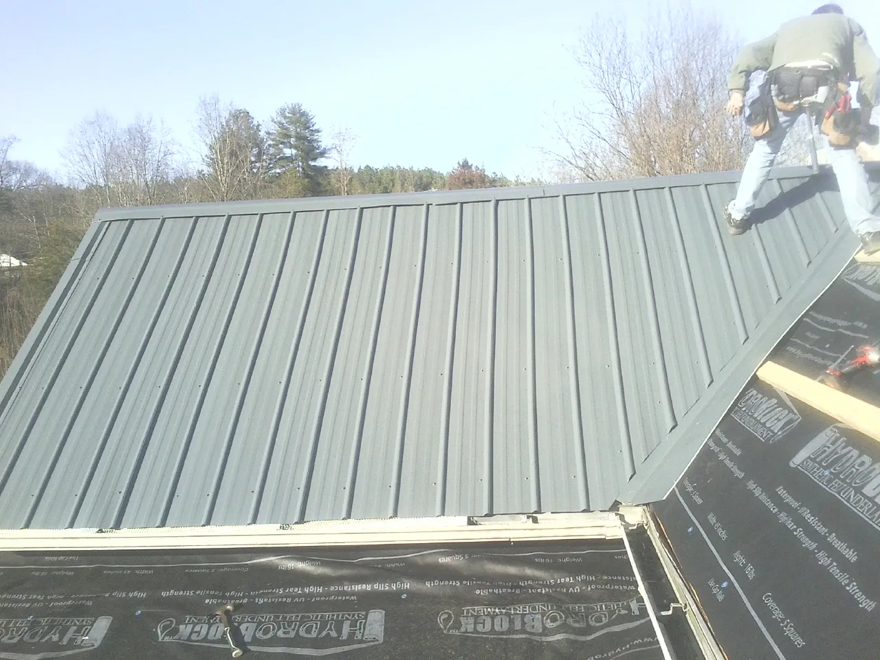Crew installing dark green standing seam panels over synthetic underlayment in Willow