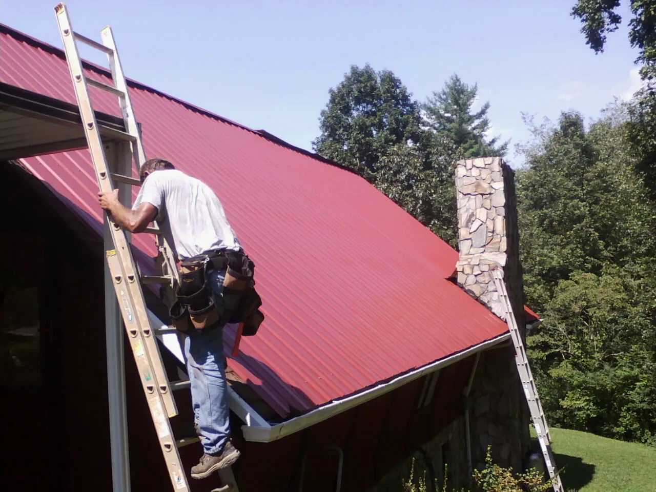 Roofer on a ladder beside a red metal roof and stone chimney — Soffit & Fascia Repair by Roofing Brothers in Willow